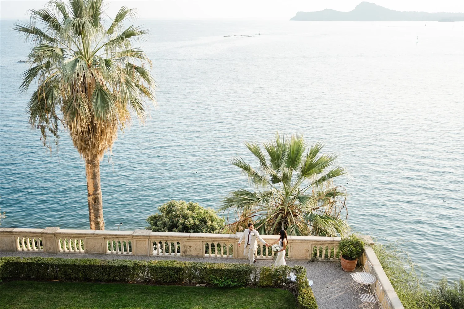 A bride and groom holding hands on a terrace overlooking the water, with palm trees and a distant landmass in the background.