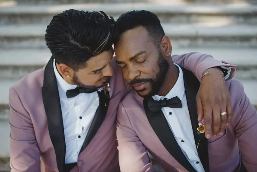 Two men in tuxedos sharing an emotional moment with their foreheads touching, sitting on outdoor steps.