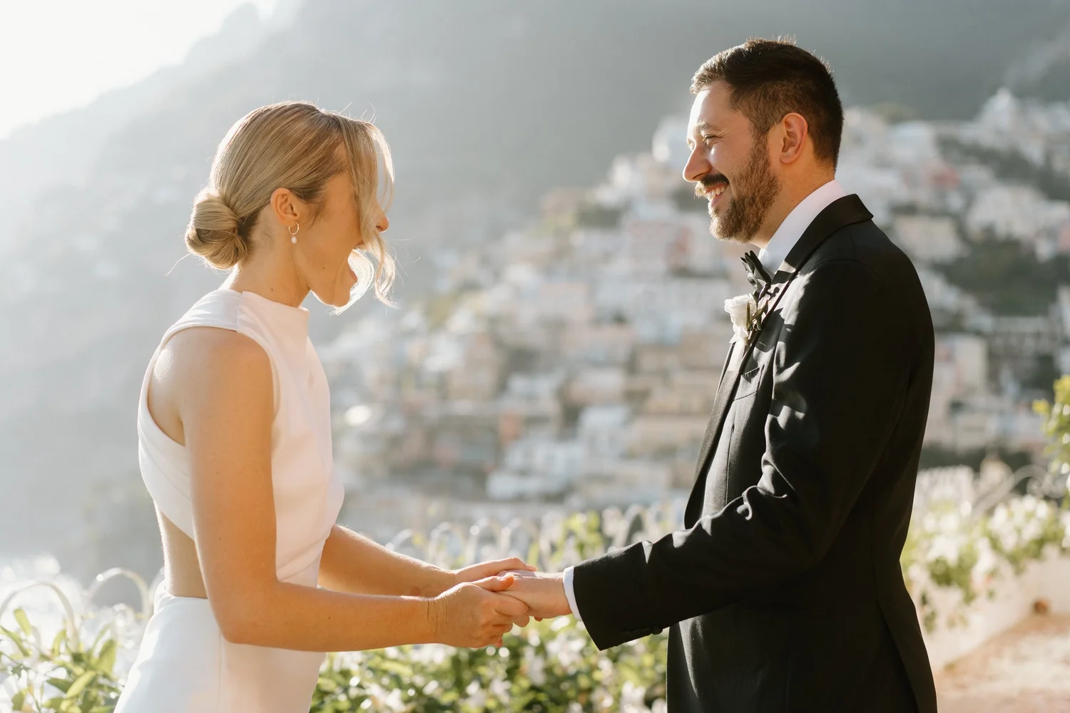 A bride and groom holding hands and facing each other, smiling during their wedding ceremony outdoors with a hilly landscape in the background.