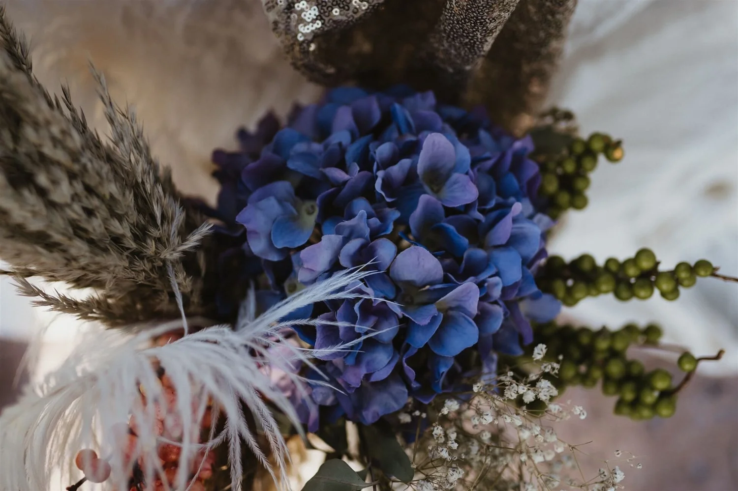 Close-up of purple hydrangea flowers, green berries, white baby’s breath, and dried pampas grass in a floral arrangement.