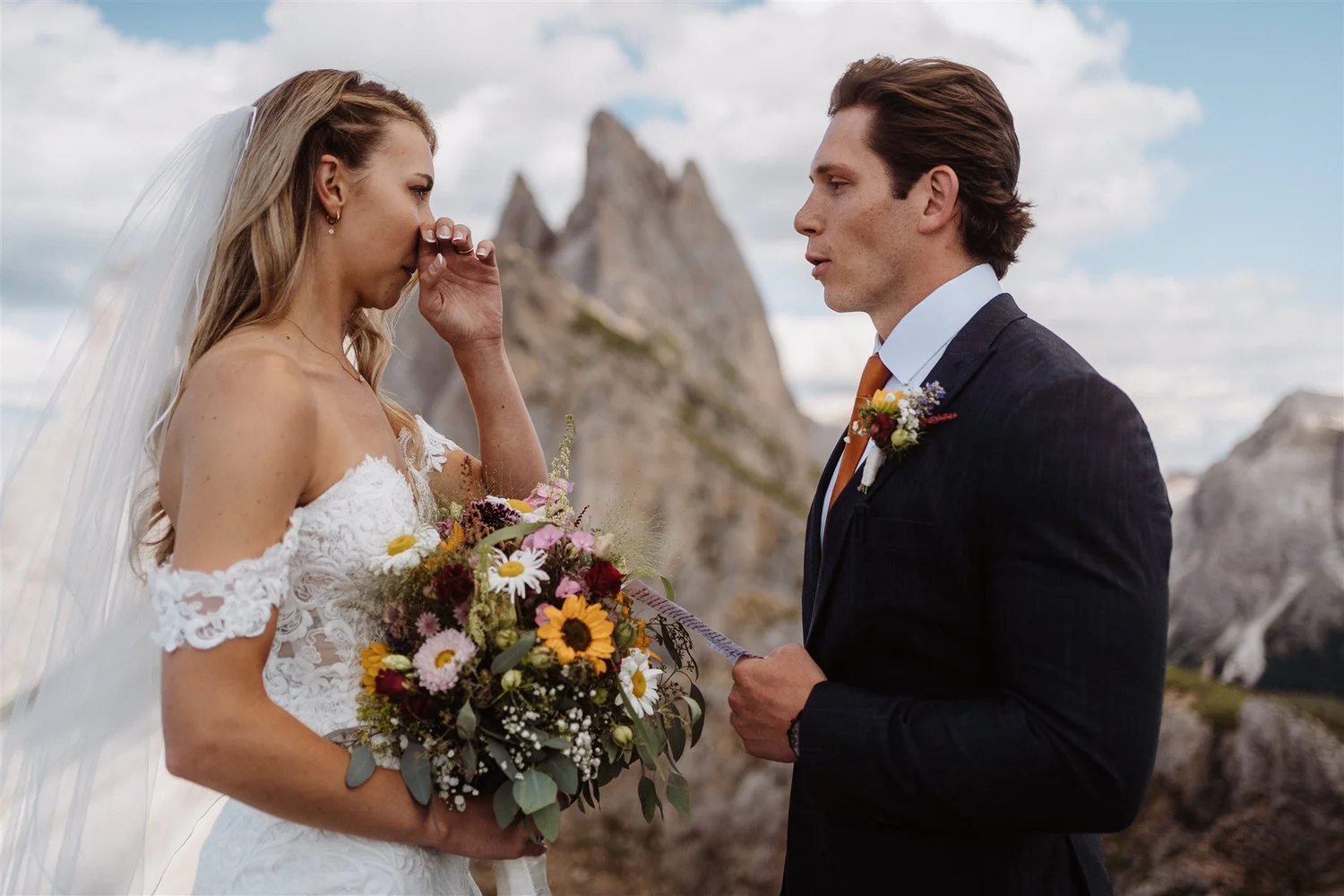 A bride and groom exchanging vows outdoors with rocky formations in the background. The bride is holding a colorful bouquet and appears emotional, while the groom is dressed in a suit with a boutonniere.