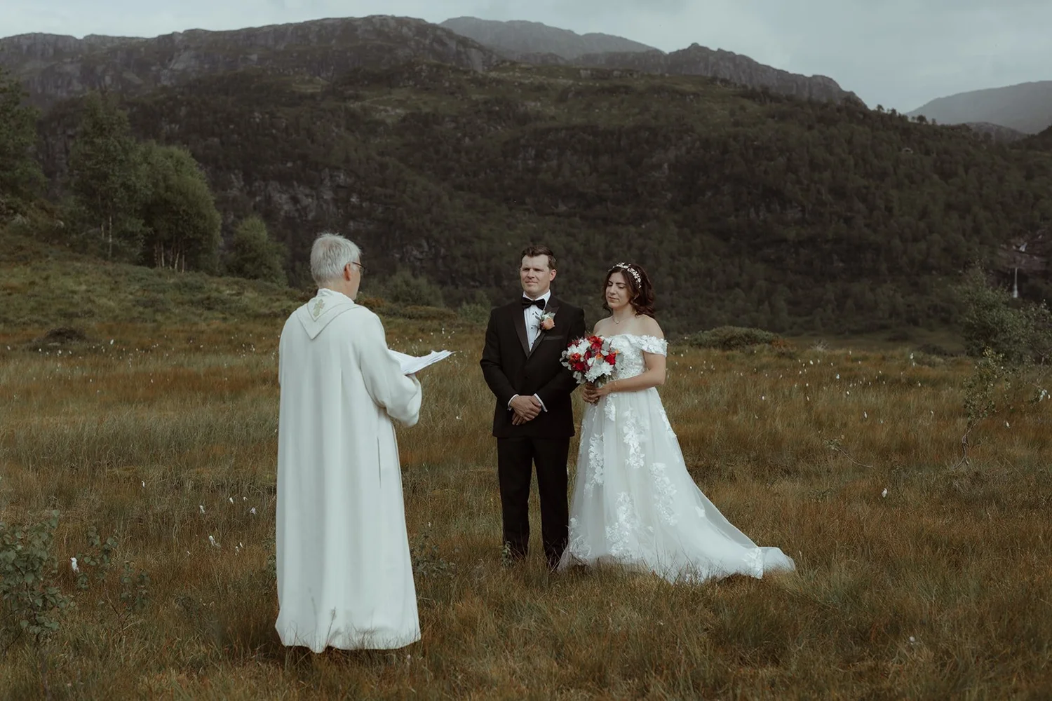 A wedding ceremony taking place outdoors in a grassy field with mountains in the background, featuring a bride in a white wedding dress holding a bouquet and a groom in a black suit with a bow tie, standing before an officiant in white robes.