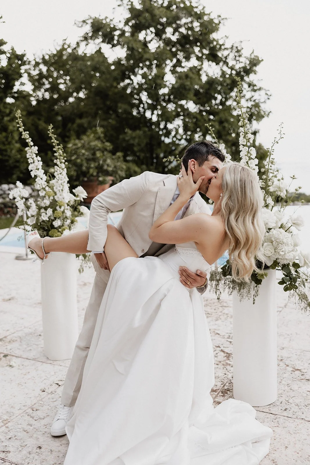 A newlywed couple sharing a kiss during their outdoor wedding ceremony, with the groom holding the bride in his arms, surrounded by white flowers and greenery.