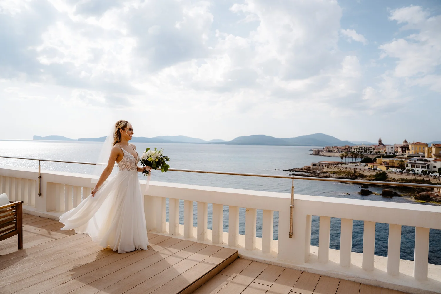 A bride in a white wedding dress holding a bouquet of white flowers on a balcony overlooking the water and a distant coastal town under a cloudy sky.