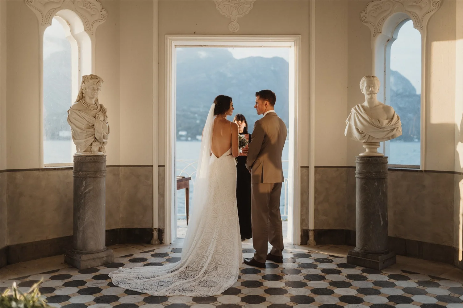 A bride and groom exchanging vows in a church with large windows and mountain views, flanked by classical statues.