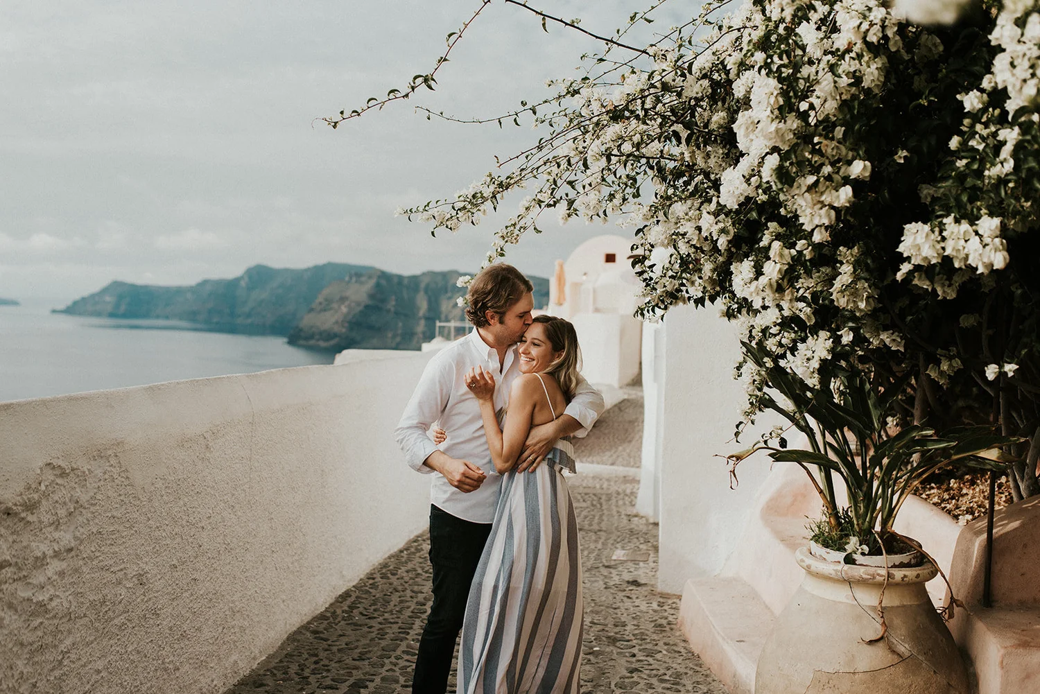A couple hugging and smiling on a terrace overlooking the ocean and cliffs, with white flowering plants and a white building in the background.