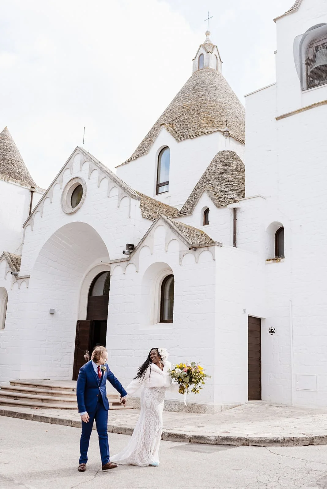 A couple in wedding attire holding hands outside a white church with a conical roof and arched windows, the bride holding a bouquet of flowers, during daytime.