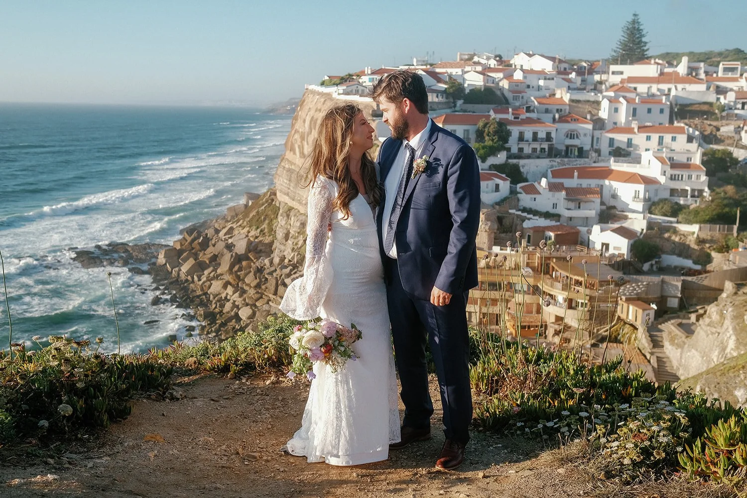 A bride and groom stand close on a cliff overlooking the ocean, with a coastal town in the background. The bride is in a white lace wedding dress holding a bouquet, and the groom is in a dark blue suit.