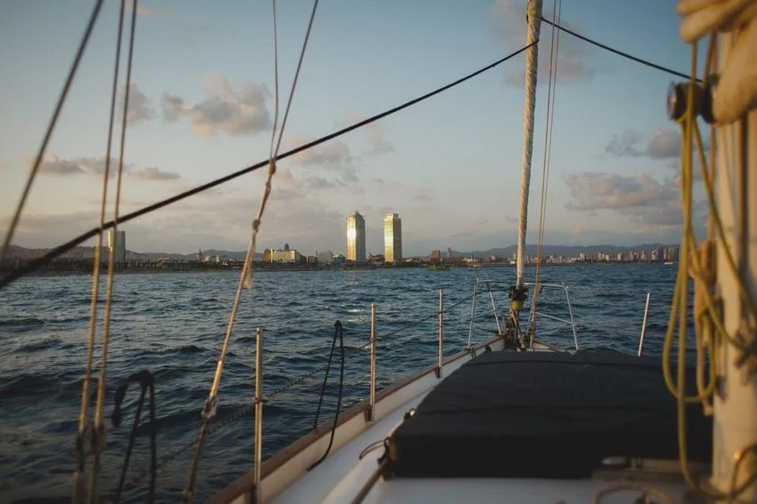 View from a boat showing water in the foreground and a city skyline with tall buildings in the background, taken during sunset with clouds in the sky.