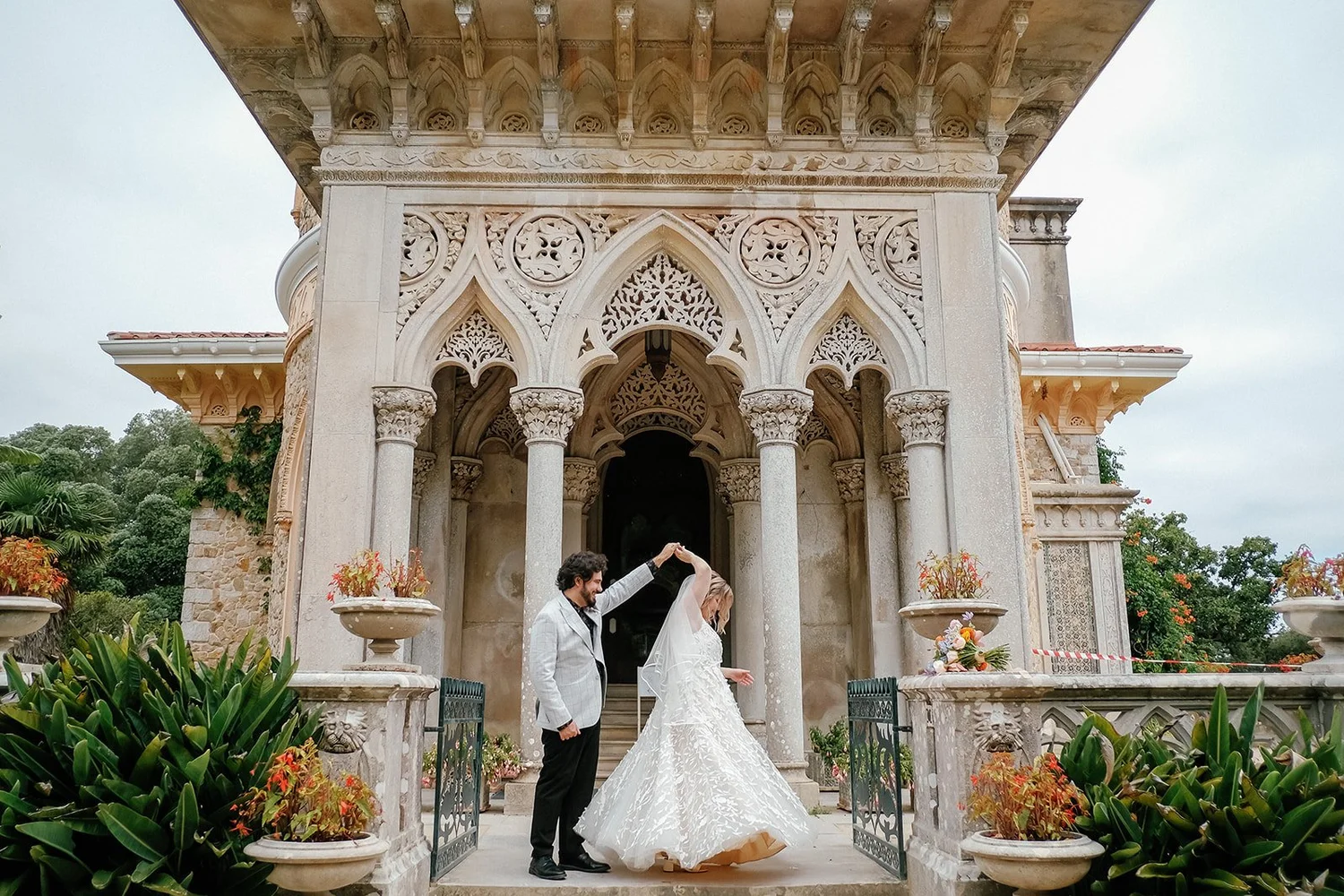 A bride and groom dancing in front of an ornate stone building with gothic arches, columns, and decorative carvings, surrounded by potted plants and greenery.