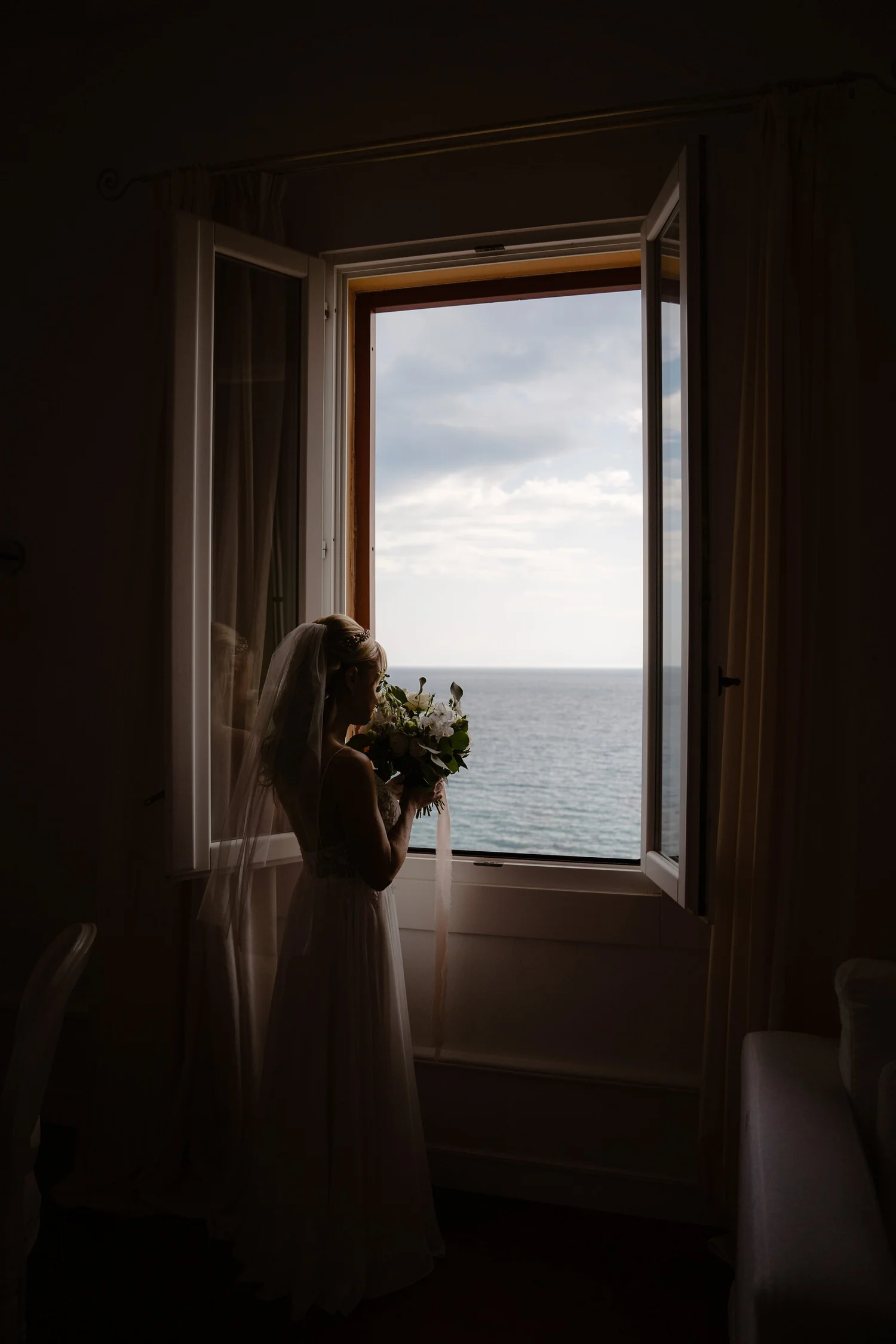 A woman in a wedding dress and veil holding a bouquet of flowers, standing in a dark room in front of an open window with light streaming in, overlooking the ocean and sky.