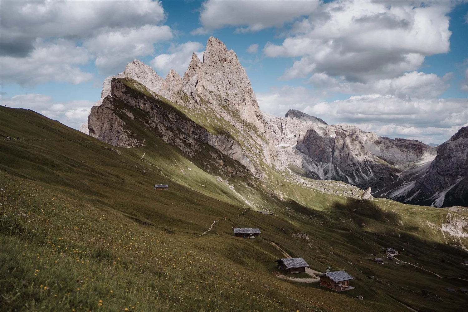 Mountain landscape with rocky peaks, green slopes, scattered small houses, and a sky filled with clouds.