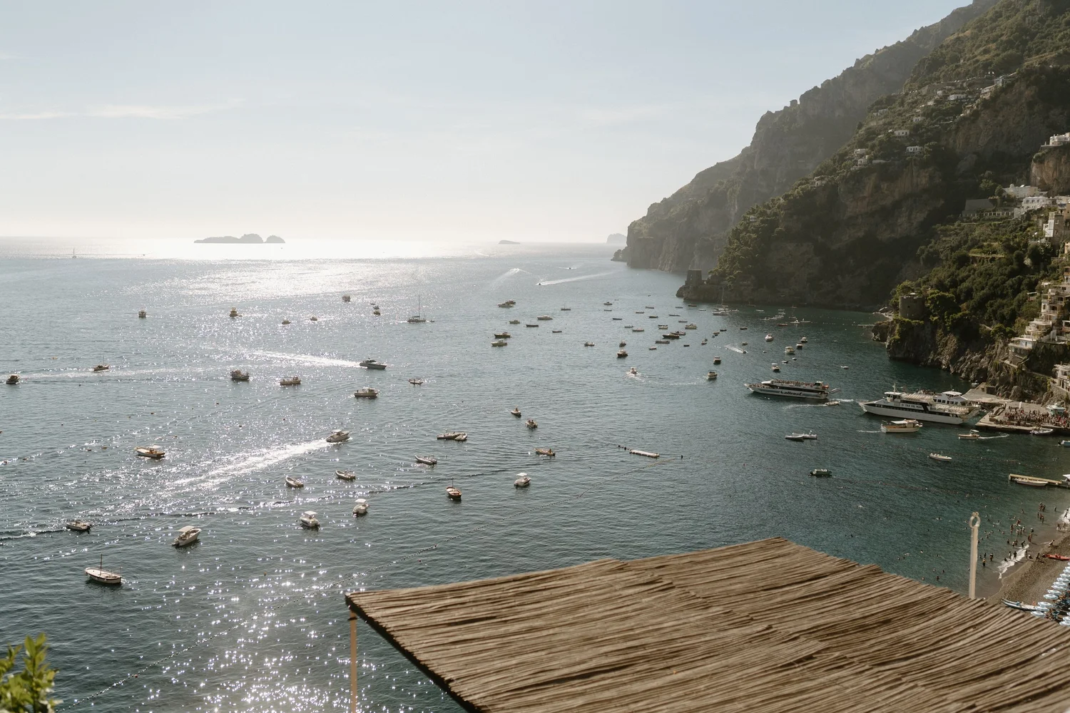 A scenic view of a coastal area with numerous boats floating on the water, cliffs with greenery and houses on the right, and a hazy sky and islands in the distance.