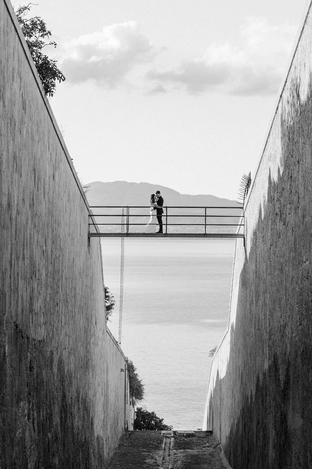 A black and white photo of a couple kissing on a small bridge between two high walls with a mountain and sky in the background.