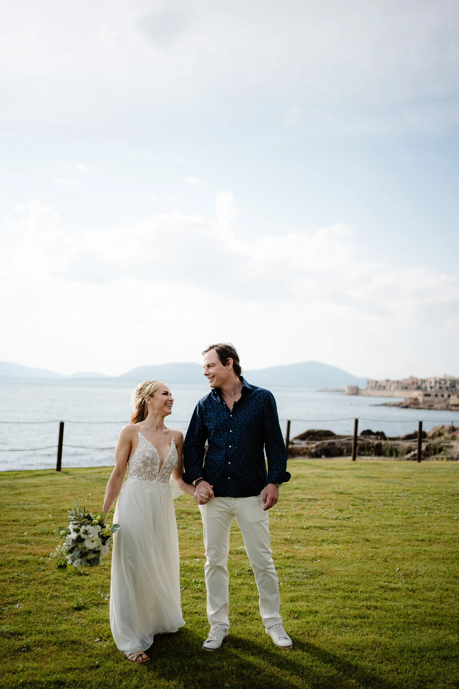 A happy couple, dressed in wedding attire, holding hands and walking outdoors on a grassy area near the water with a rocky shoreline, buildings, and mountains in the background, under a partly cloudy sky.