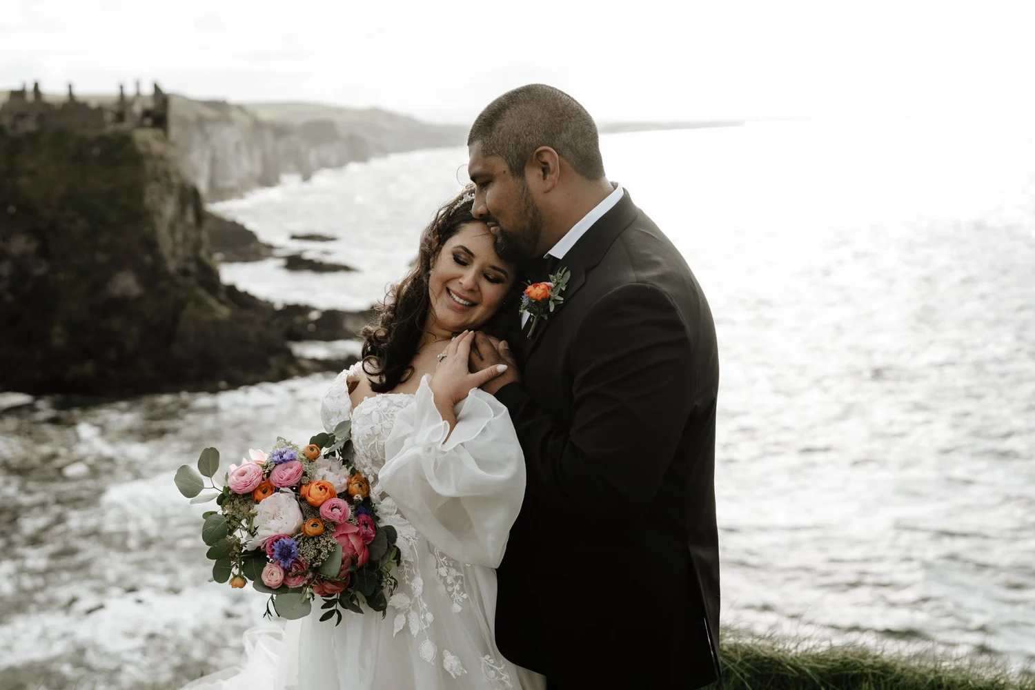 A newlywed couple hugging by the ocean on a cloudy day, with cliffs in the background. The bride is holding a bouquet of colorful flowers and smiling, while the groom is gently resting his forehead on her head.