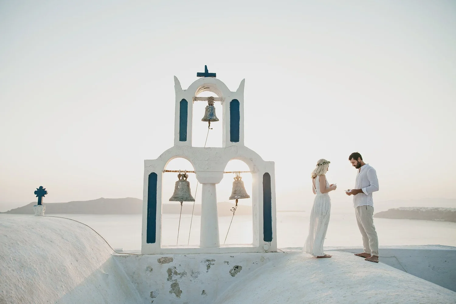 A couple dressed in white wedding attire standing outdoors, exchanging vows in front of a white church bell tower with three bells, against a clear sky and distant mountains.