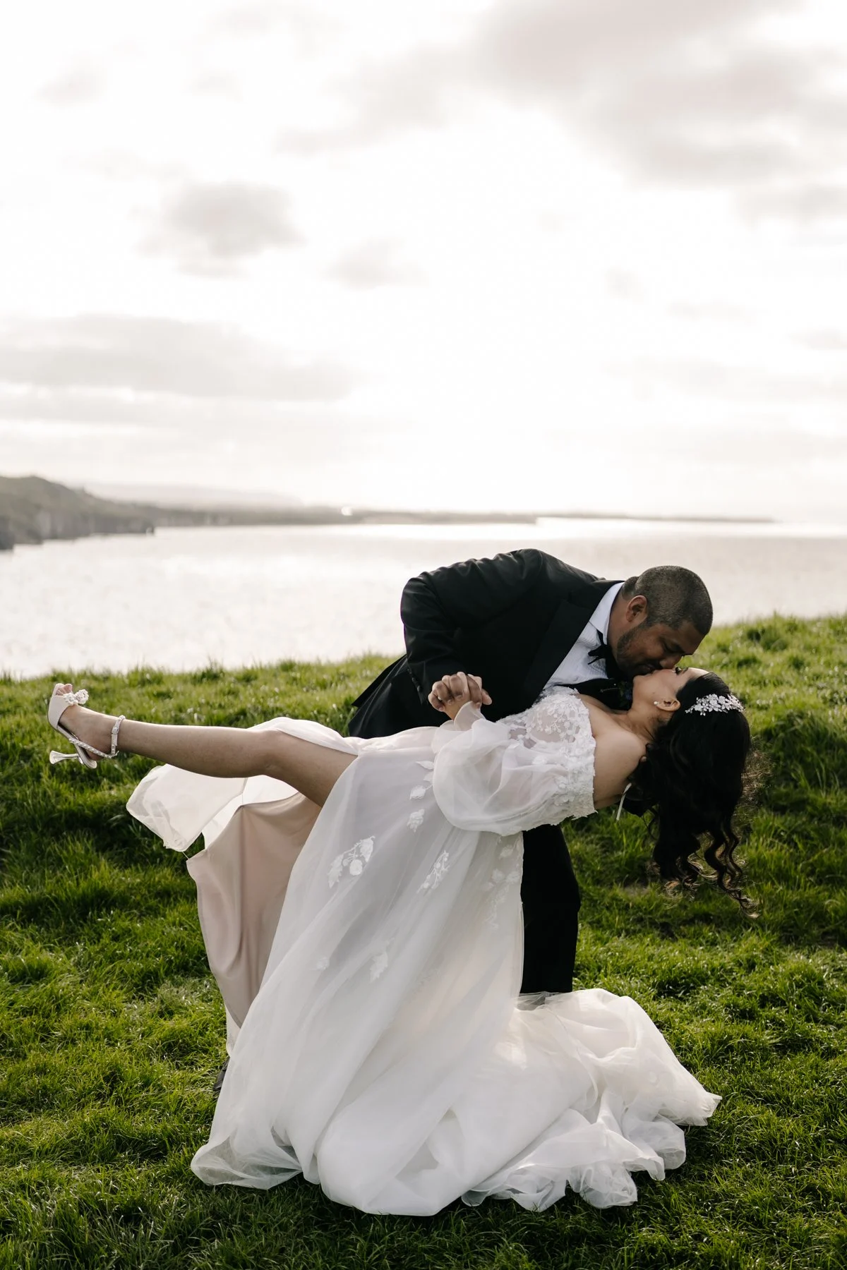 A bride and groom sharing a kiss outdoors by a lake; the groom is holding the bride in a dip, both dressed in wedding attire, with the bride wearing a white dress and the groom in a black tuxedo.
