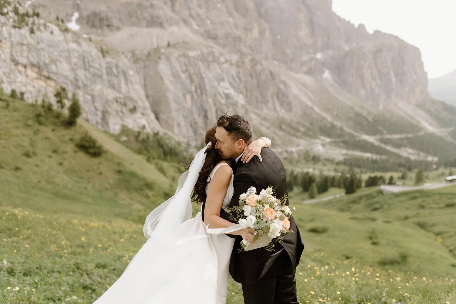 A bride and groom hugging outdoors with mountainous landscape in the background, the bride holding a bouquet of flowers.