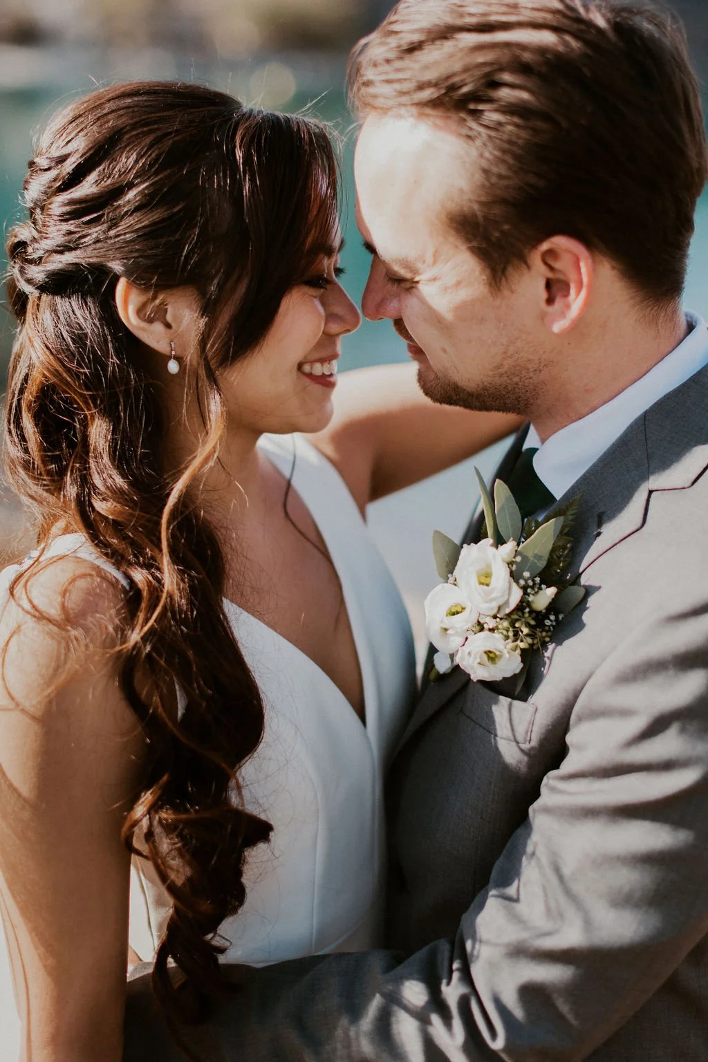 A couple with their foreheads touching, smiling during a wedding photo, outdoors in sunlight, with the woman in a white dress and the man in a suit with a boutonniere.