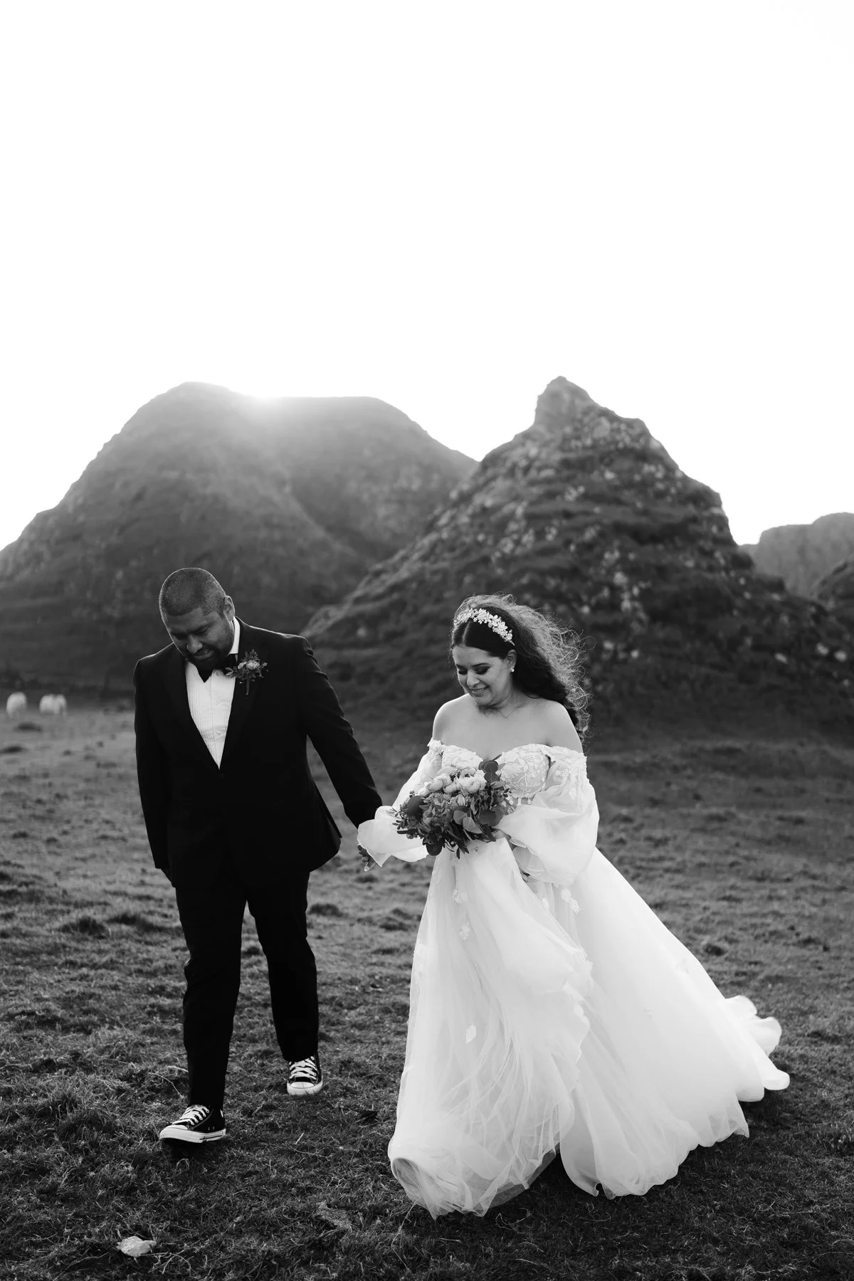 Black and white photo of a bride and groom walking in a grassy field with mountains in the background. The bride is wearing an off-shoulder wedding dress and holding a bouquet, while the groom is in a suit with sneakers, holding her hand and looking