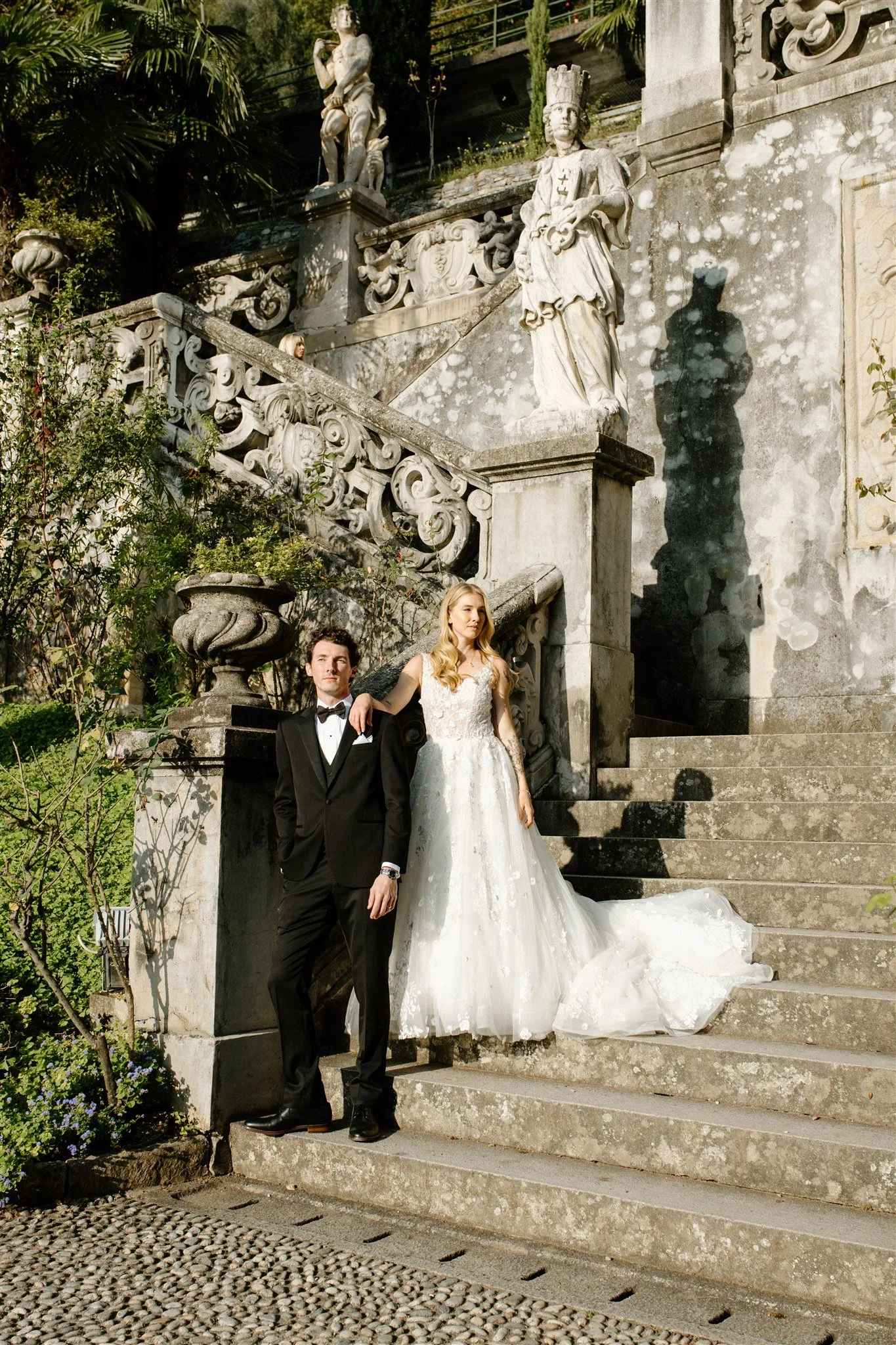 A bride and groom pose on stone stairs in front of classical sculptures, with lush greenery surrounding the scene.