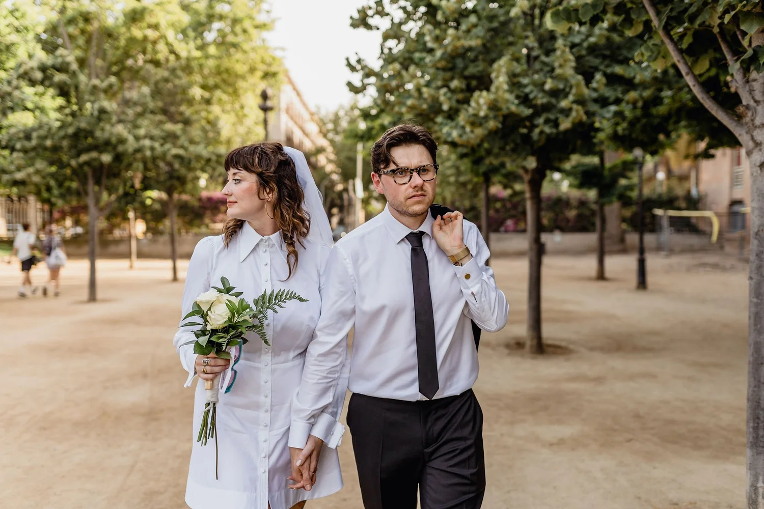 A bride and groom walking outdoors on a sunny day, holding hands, with the bride holding a bouquet of white flowers and greenery. The bride wears a white dress with a veil, and the groom wears a white shirt, black tie, and glasses. They are surrounde