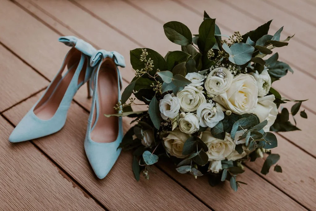 A pair of light blue high heel shoes with bow ties, placed next to a bouquet of white roses and greenery on a wooden floor.