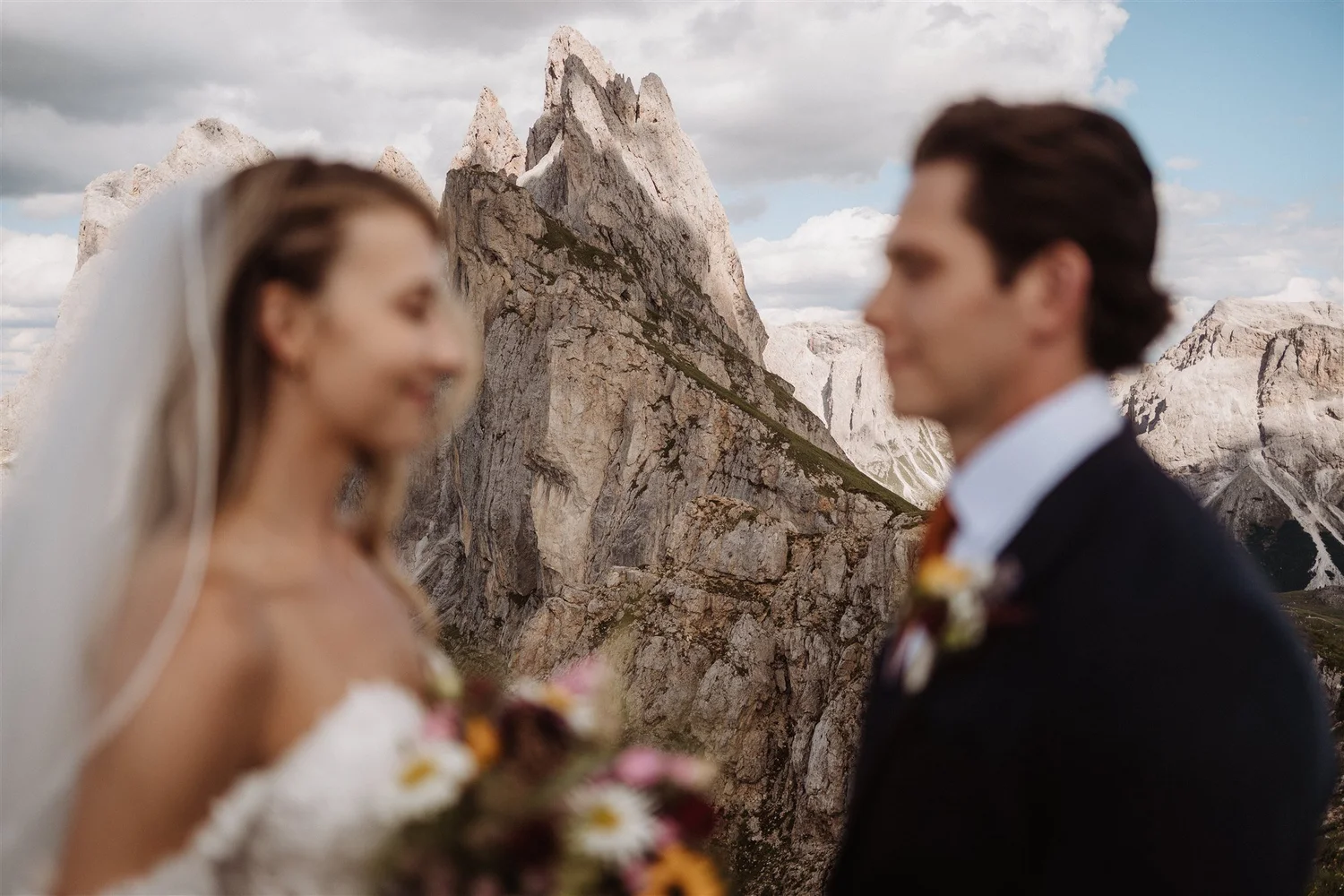 Blurred image of a bride and groom facing each other outdoors with mountain peaks in the background.