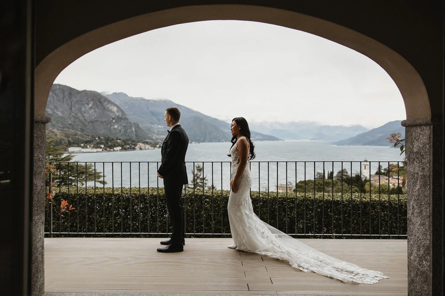 A bride and groom standing on a wooden deck under an archway overlooking a lake and mountains.