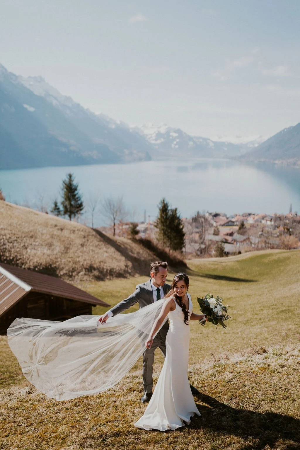 A bride and groom in wedding attire standing on a grassy hillside with a lake and mountains in the background, the bride holding a bouquet and the groom gently pulling her veil.