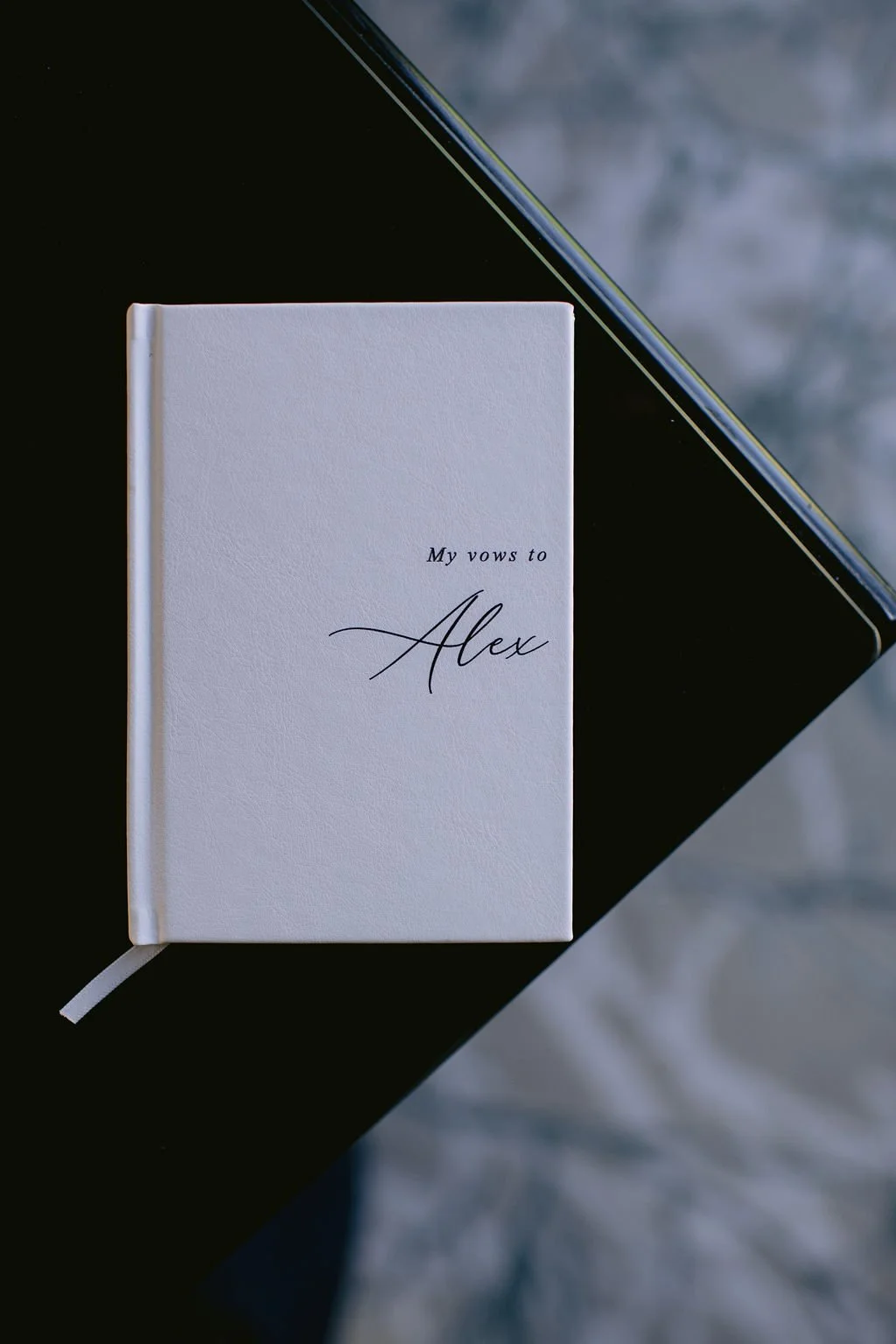 A white notebook with the words 'My vows to Alex' written on the cover, placed on a black surface with a diagonal edge, with a blurred background.