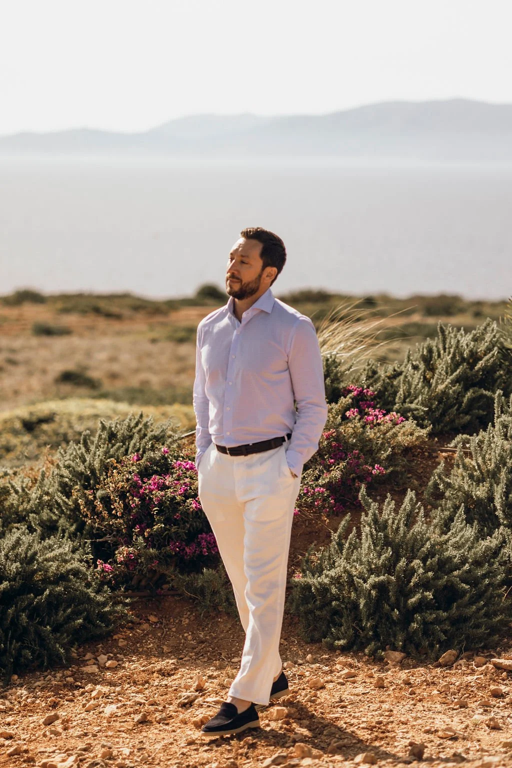 A man in a light pink dress shirt and white pants walking outdoors in a desert landscape with shrubs and pink flowers, with mountains and a lake in the background.
