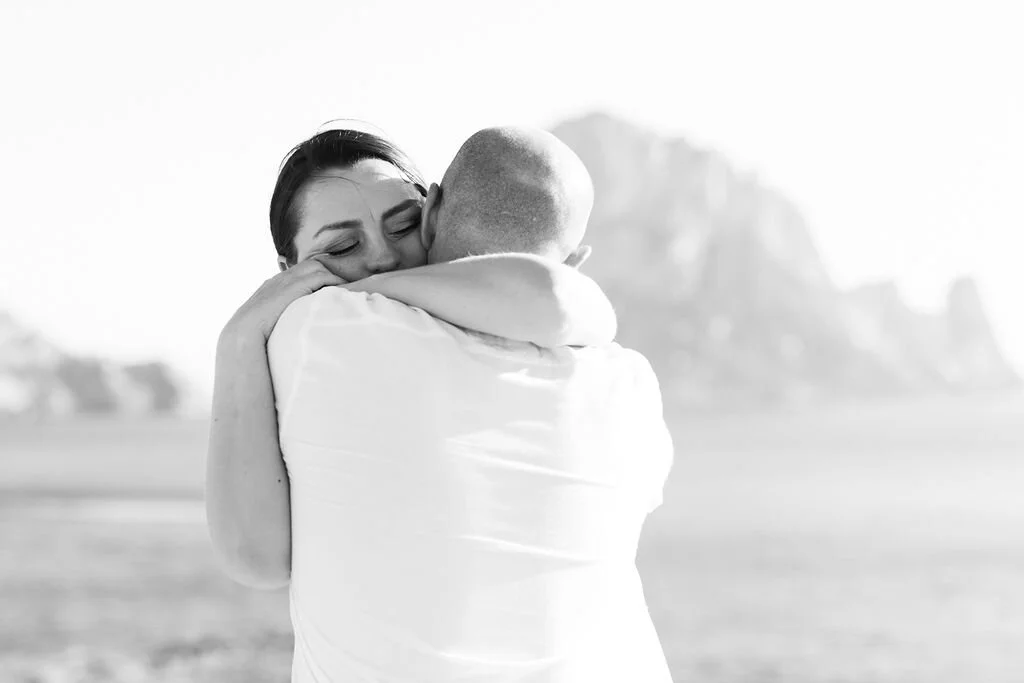 A woman and a man hugging on a beach, with the woman smiling, and a large rock formation in the background.