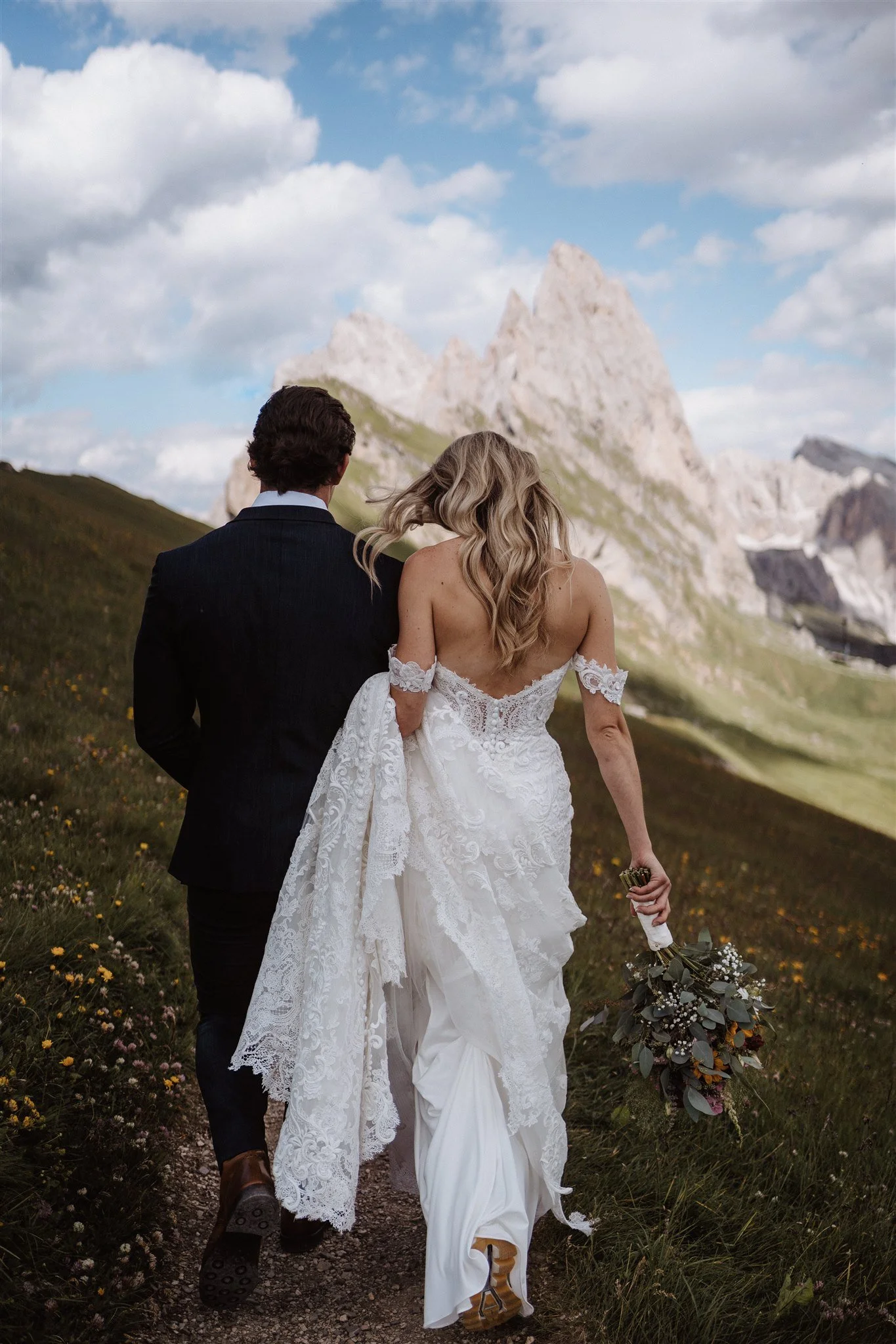 A bride and groom walking hand in hand on a mountain trail with mountains and a partly cloudy sky in the background, the bride holding a bouquet of flowers.