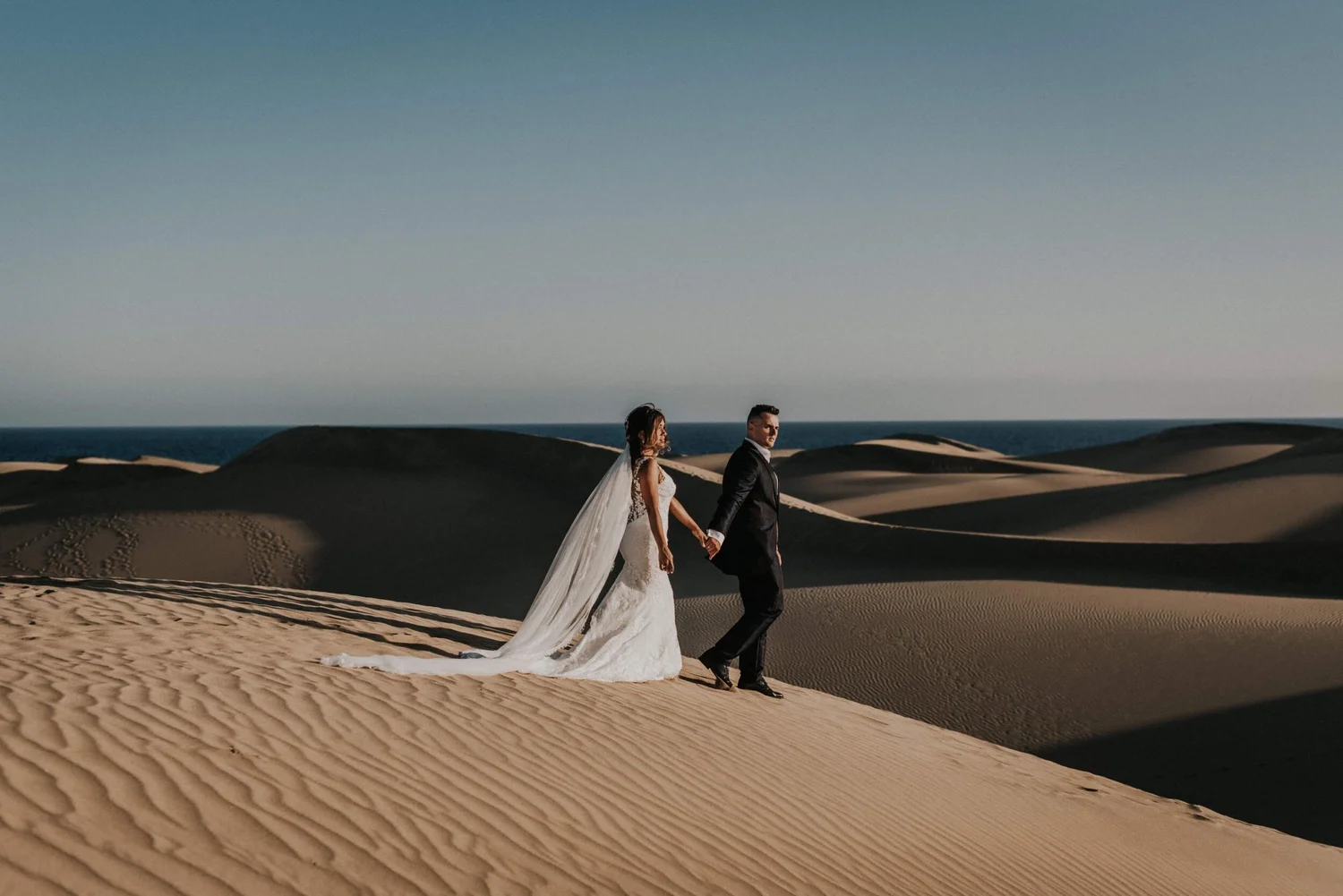 A bride and groom walking hand in hand through sand dunes in a desert landscape during daytime.