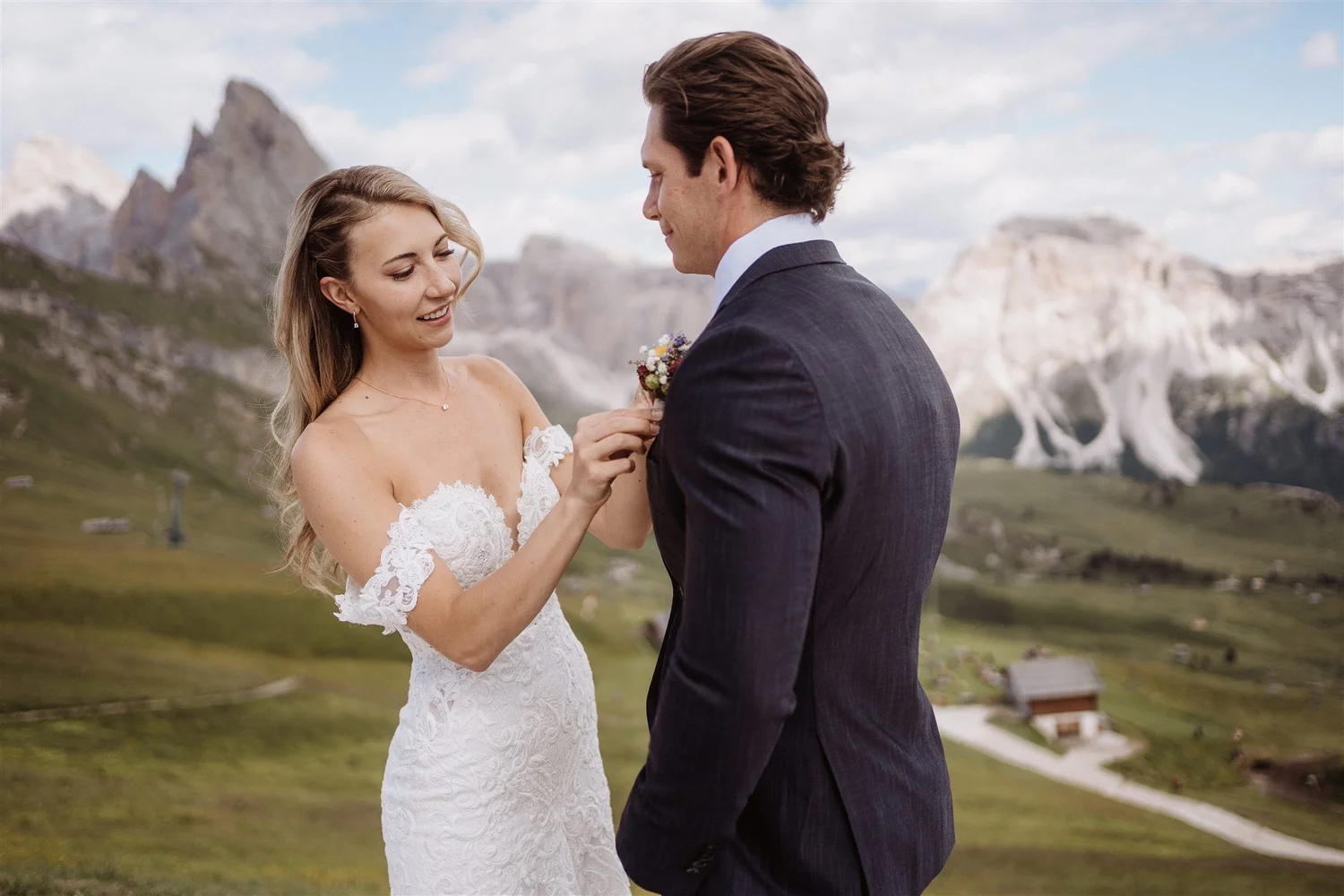 A bride and groom are standing outdoors in a mountainous landscape. The bride, wearing a white lace wedding dress, is smiling as she holds a small bouquet, and the groom, in a dark suit, looks at her with a gentle expression.