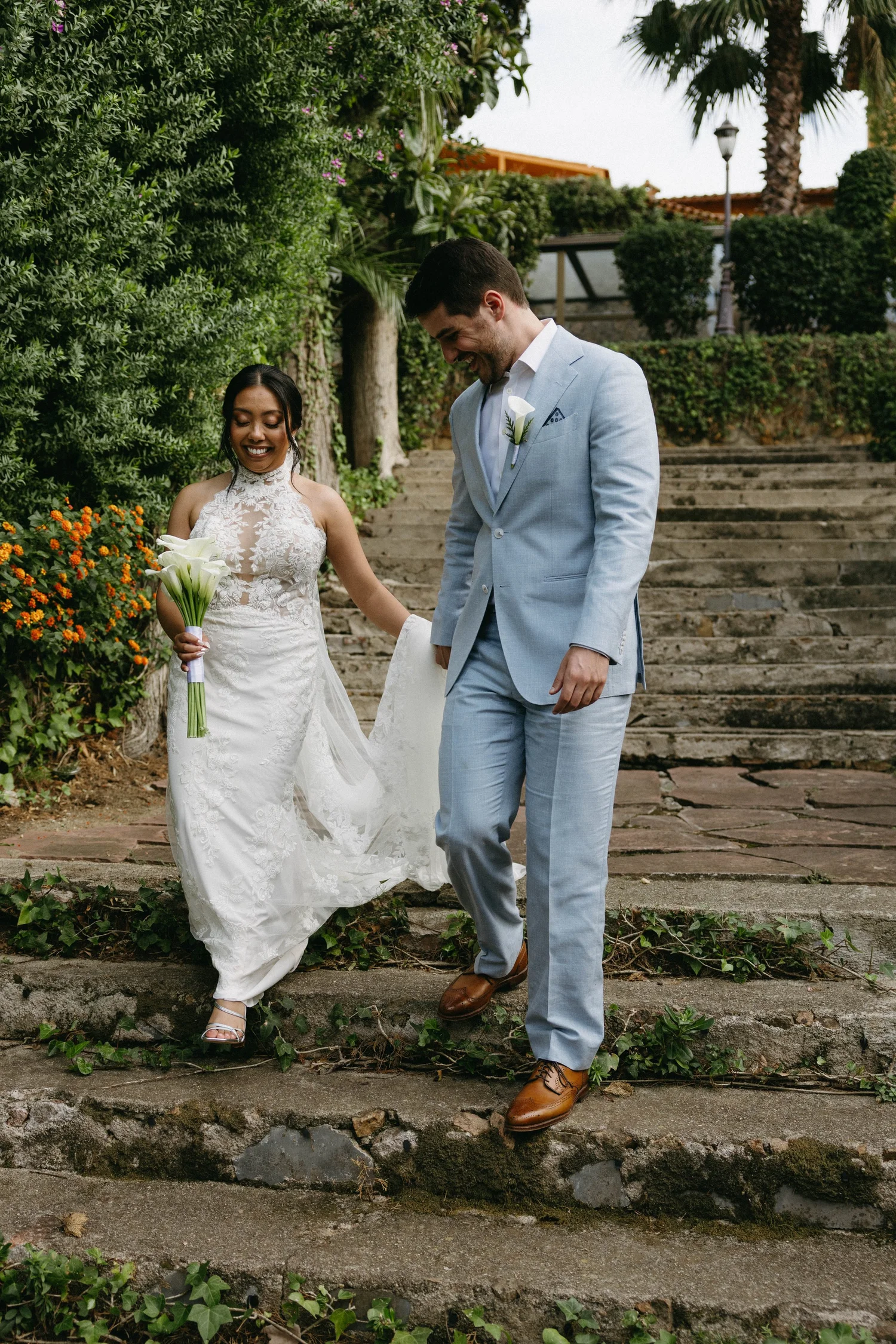 A bride and groom smiling and walking down stone steps outdoors. The bride is in a white lace wedding dress holding a bouquet of calla lilies, and the groom is in a light blue suit with a white shirt and brown shoes. They are surrounded by lush green