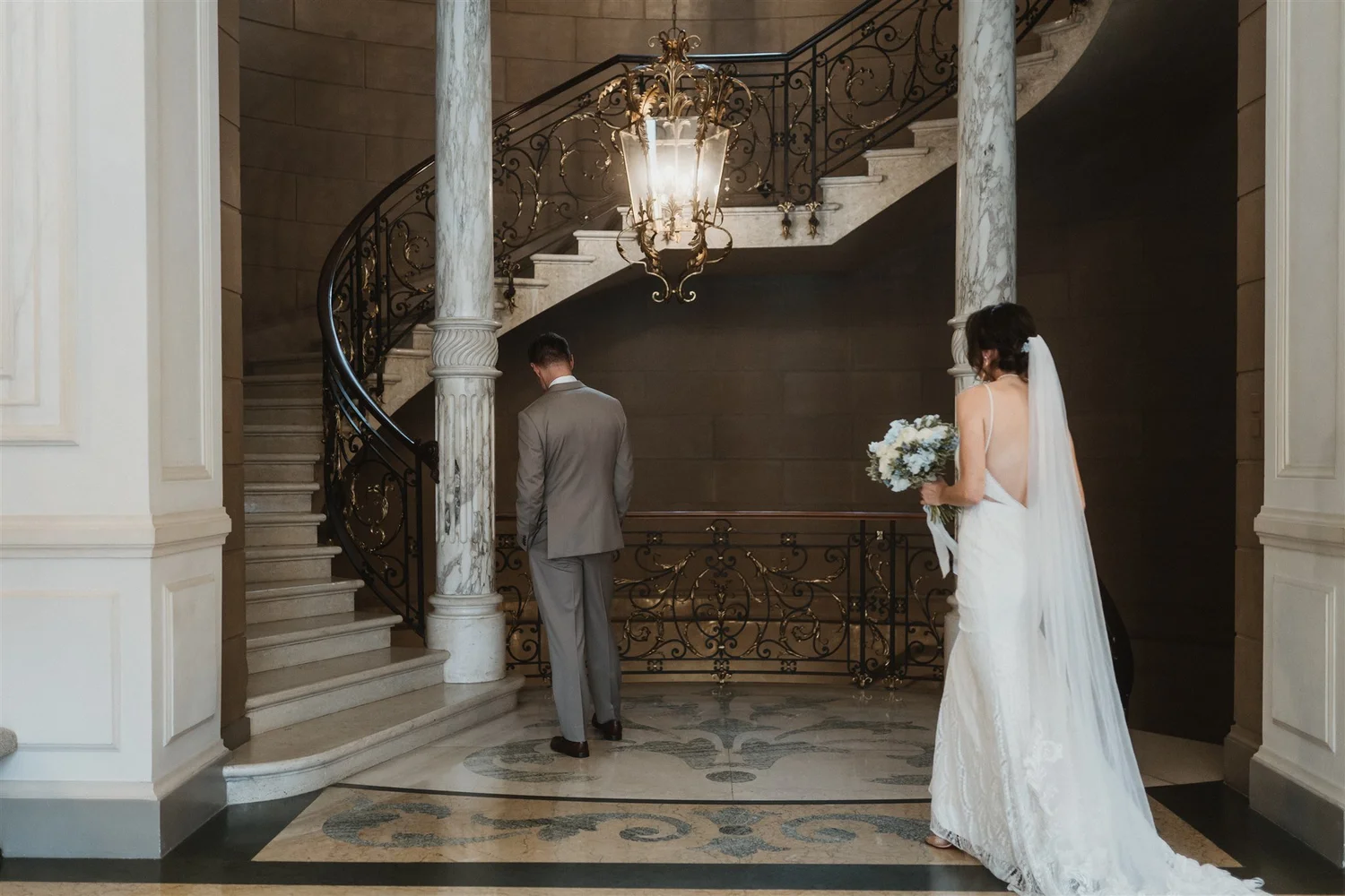 A bride and groom standing inside a grand entrance hall, with elegant staircase, marble columns, and ornate chandelier. The bride is holding a bouquet, and the groom is dressed in a gray suit, facing away from the camera.