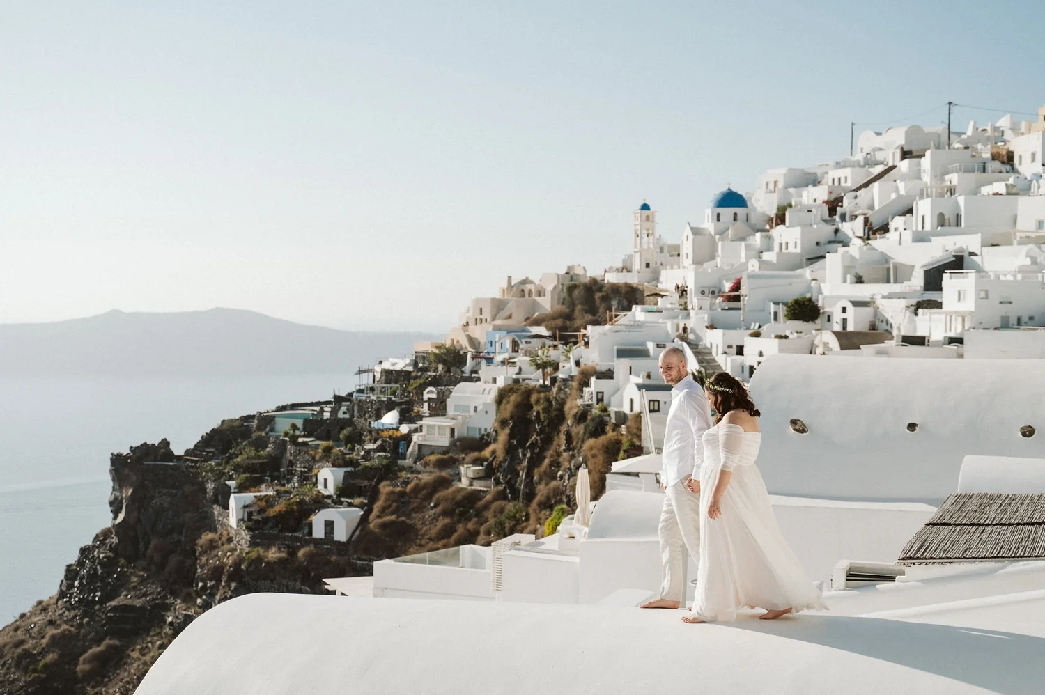 A couple dressed in white wedding attire standing barefoot on a white platform, overlooking the iconic cliffside buildings of Santorini, Greece, with blue-domed churches and caldera in the background.