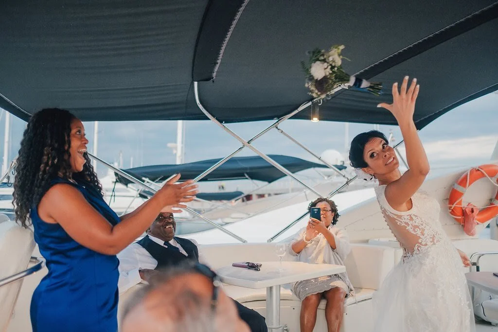 Women celebrating on a yacht, with one woman in a white lace wedding dress, another in a blue dress, and others taking photos.