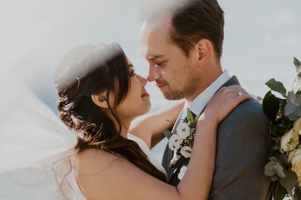 A bride and groom embrace outdoors, faces close with eyes closed, the bride's veil flowing, surrounded by natural light.