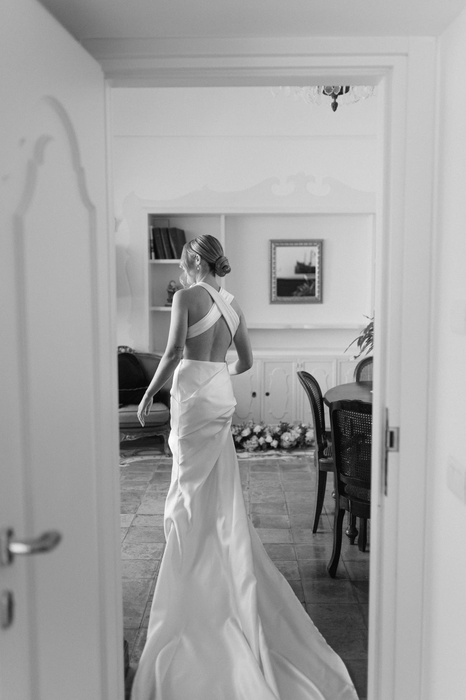 A woman in a wedding gown standing in a room, reflected through a doorway, with a chandelier overhead and a floral arrangement on the floor.