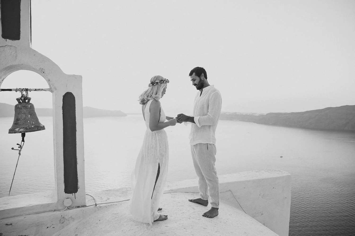 A black and white photo of a couple standing on a white-washed building overlooking the water, holding hands, with a bell and archway visible on the left, capturing a romantic moment.
