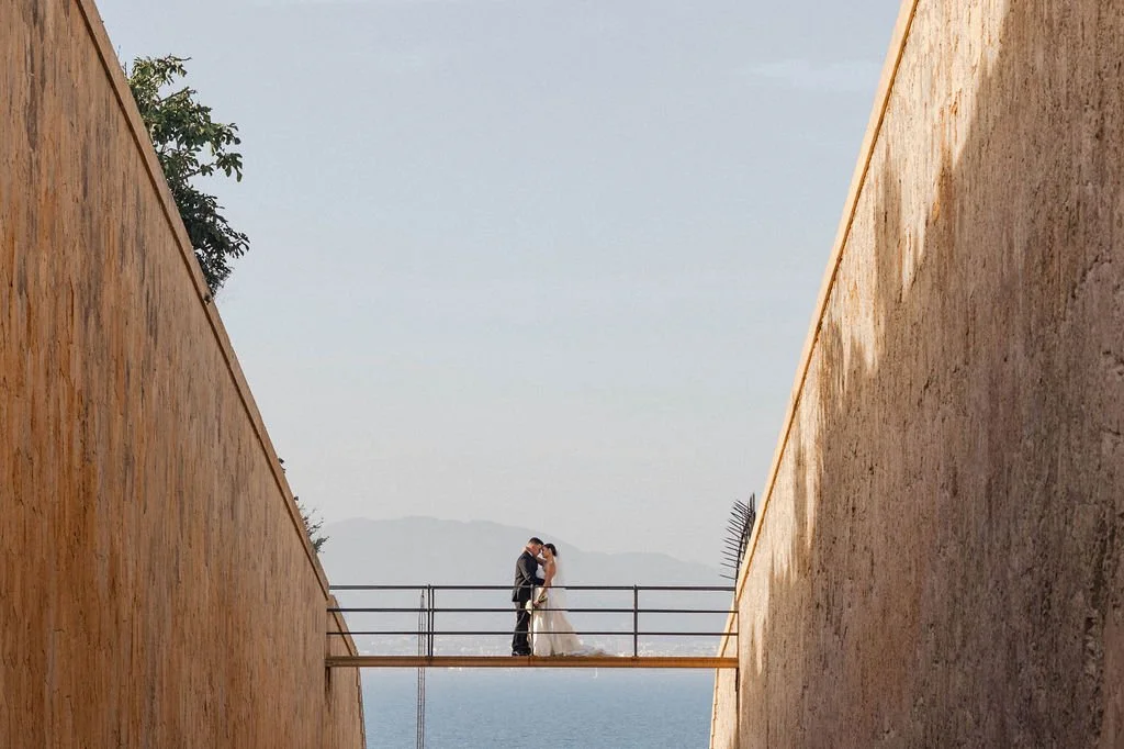 A couple in wedding attire standing on a narrow bridge between high walls, sharing a kiss with mountains and water in the background.
