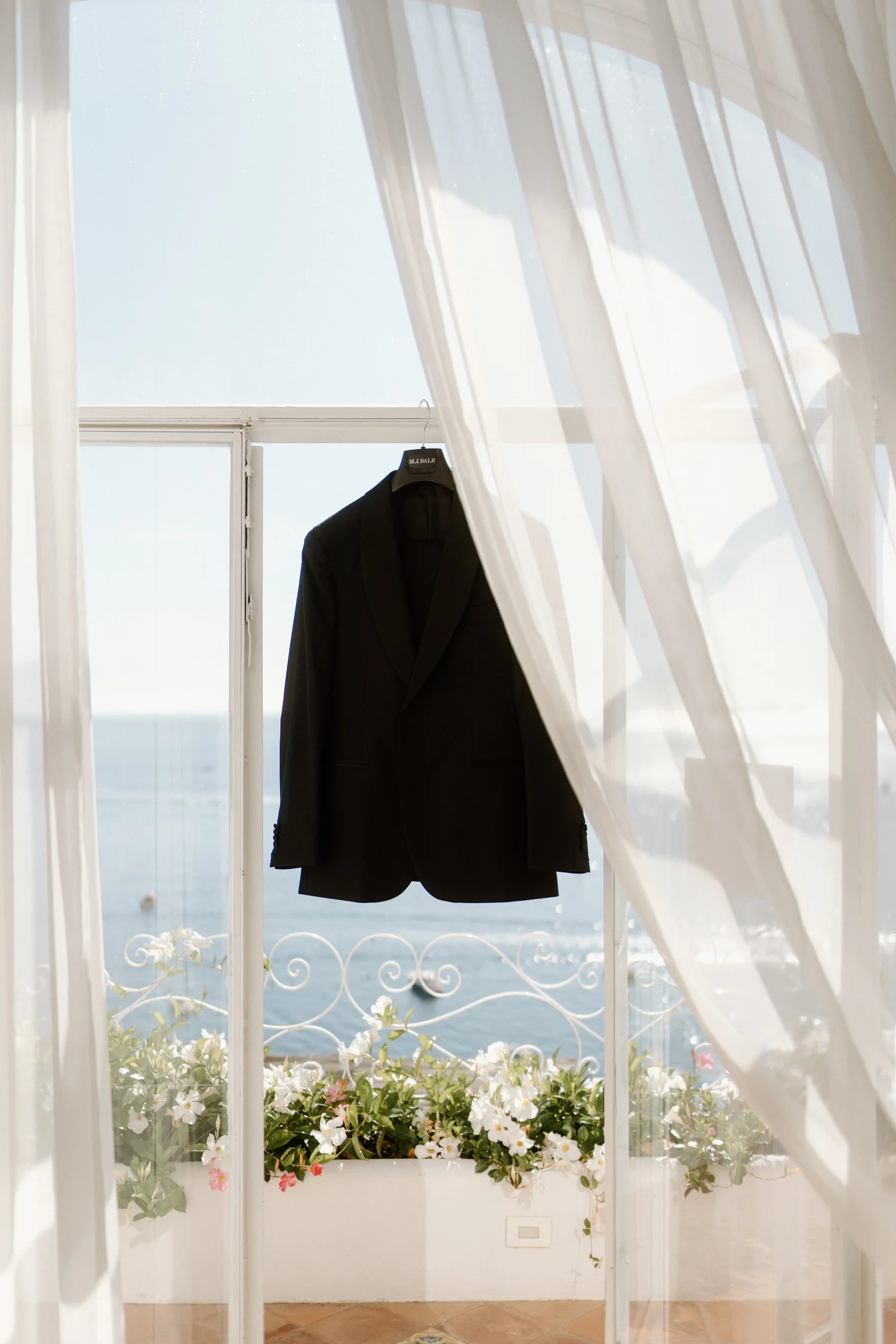 A black suit jacket hangs on a hanger on a window with white curtains. Outside the window, there is a view of the ocean and blue sky. At the bottom of the window, there are white and pink flowers in a window box.