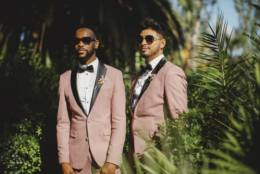 Two men in pink suits with black lapels and bow ties standing outdoors among lush greenery, both wearing sunglasses and boutonnières, with sunlight filtering through trees.