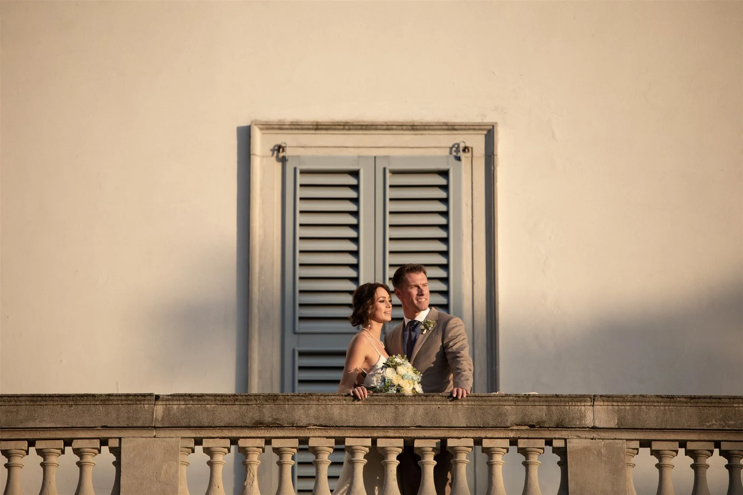 A bride and groom stand on a balcony under a window with gray shutters, with the bride holding a bouquet of white and cream flowers. The scene is lit by warm sunlight, and they are looking into the distance.