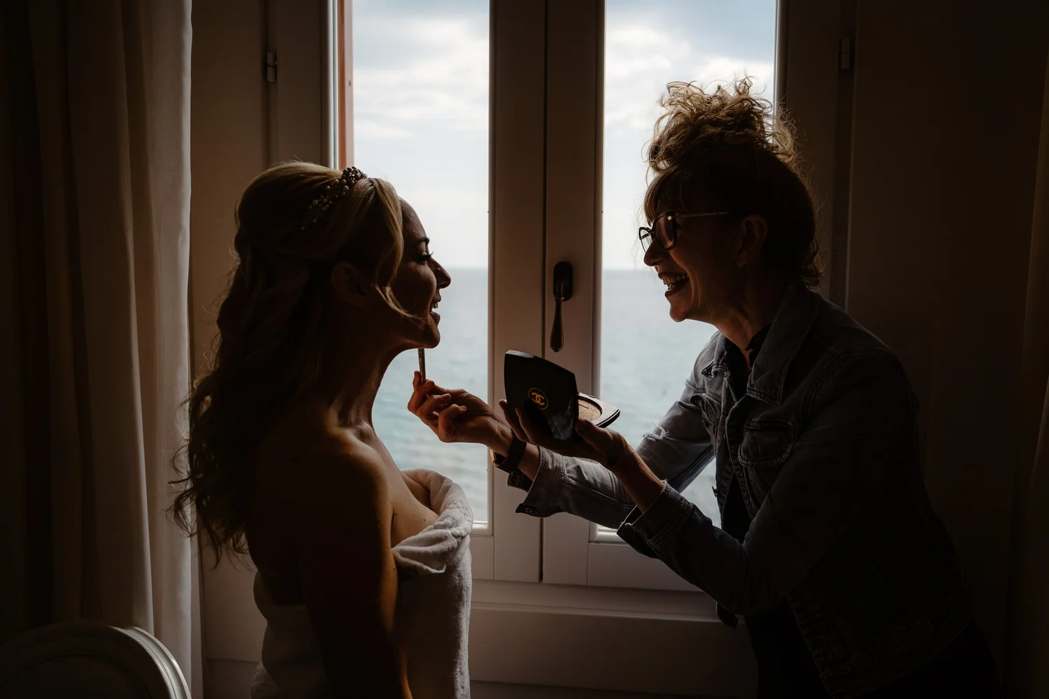 A woman with blonde hair receiving makeup application from an artist in front of a window overlooking the ocean. The woman is wrapped in a towel and has a beaded headband, while the artist is wearing glasses and a denim jacket, holding a makeup palet