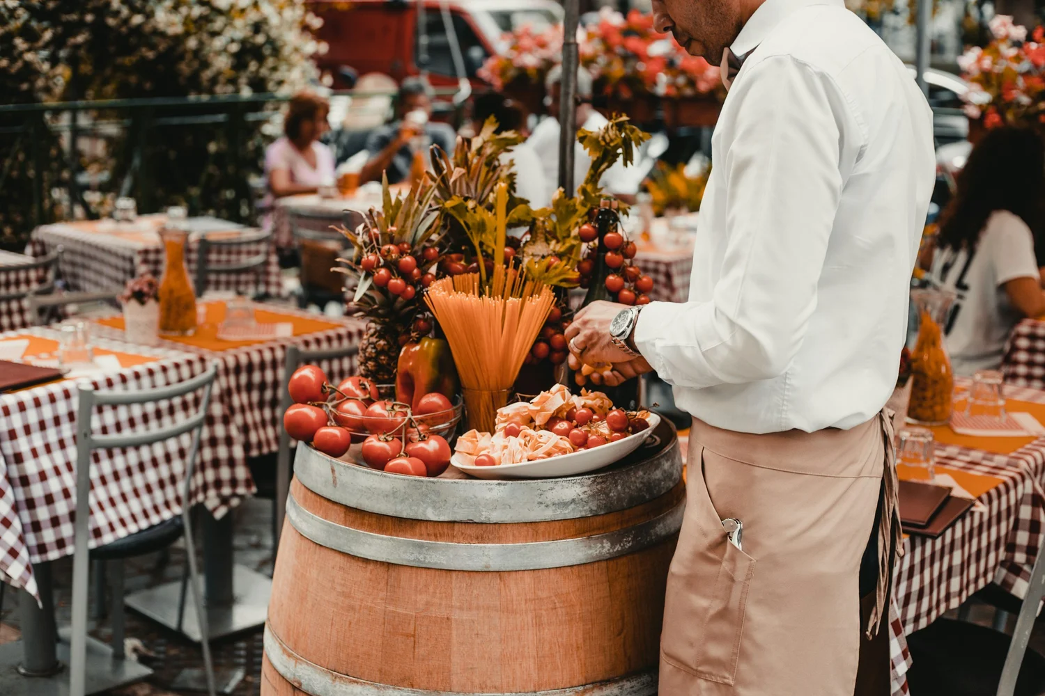 A man in a white shirt and beige apron looks at tomatoes and other produce arranged in a large barrel at an outdoor market with people sitting at tables in the background.